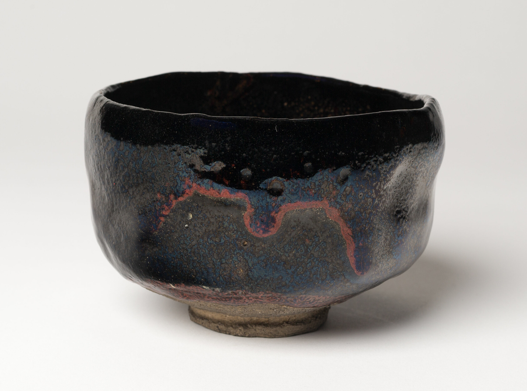 A dark glazed tea bowl with an uneven rim and blue gray and rusty red patches on a pale background.