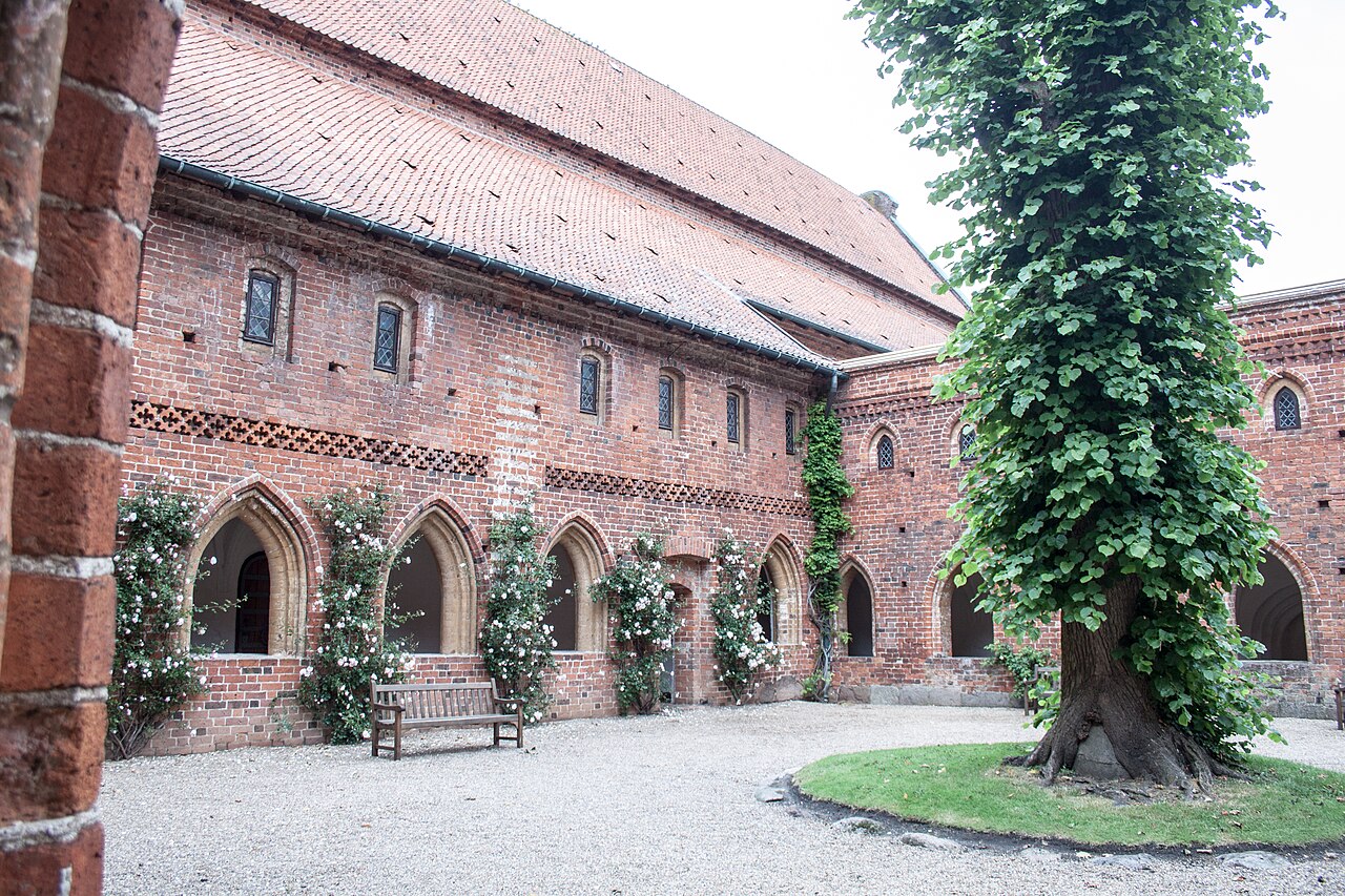 A brick cloister with pointed arches and a bench around a gravel courtyard and leafy tree.