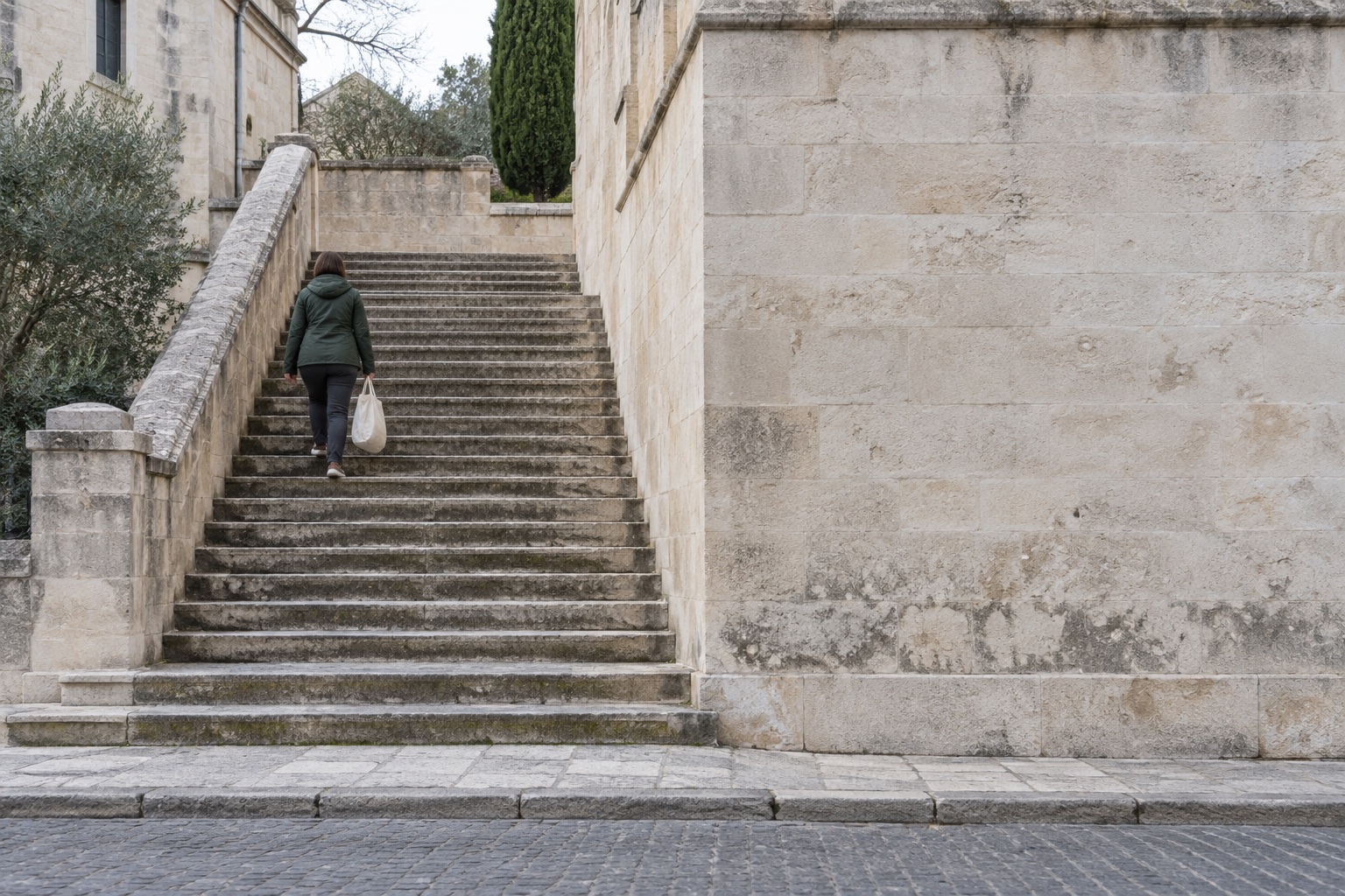 Seen from across a street, one small figure in a green jacket climbs a broad flight of pale stone steps in a town, carrying a canvas bag.