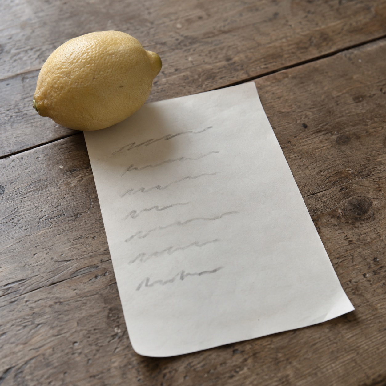 A handwritten shopping list, too faint to read, held flat under a lemon on a wooden kitchen table. Shared by Ines.