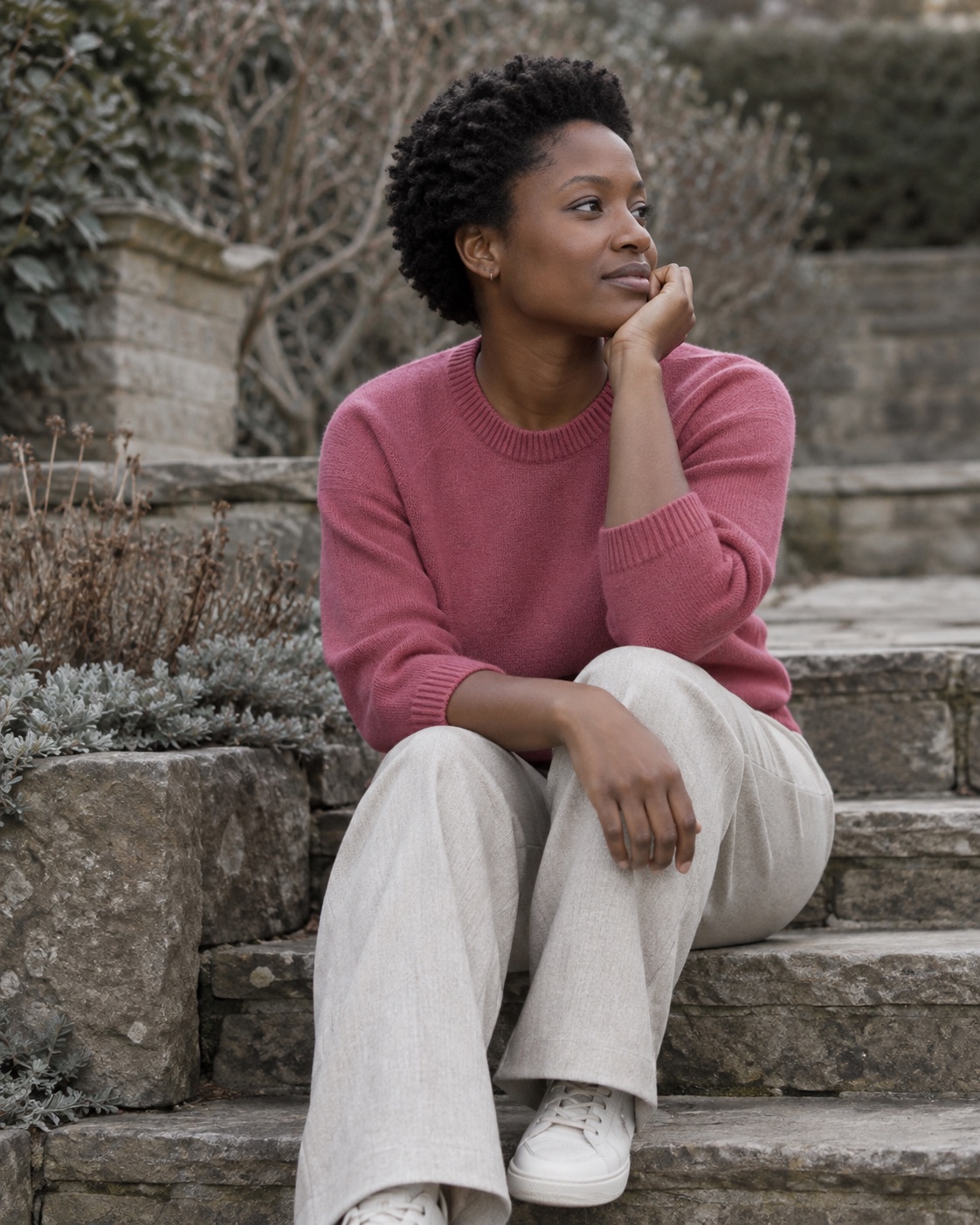 Amara, fictional, sitting on a stone garden step in her rose knit, chin resting on one hand.