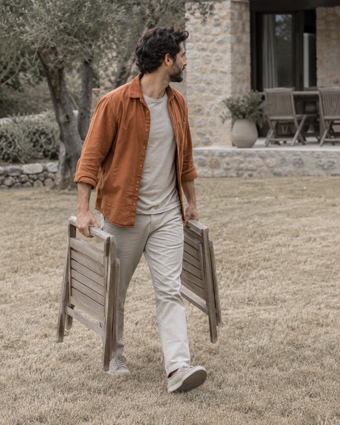 Theo, fictional, crossing a lawn in his apricot overshirt with a folded wooden chair in each hand.