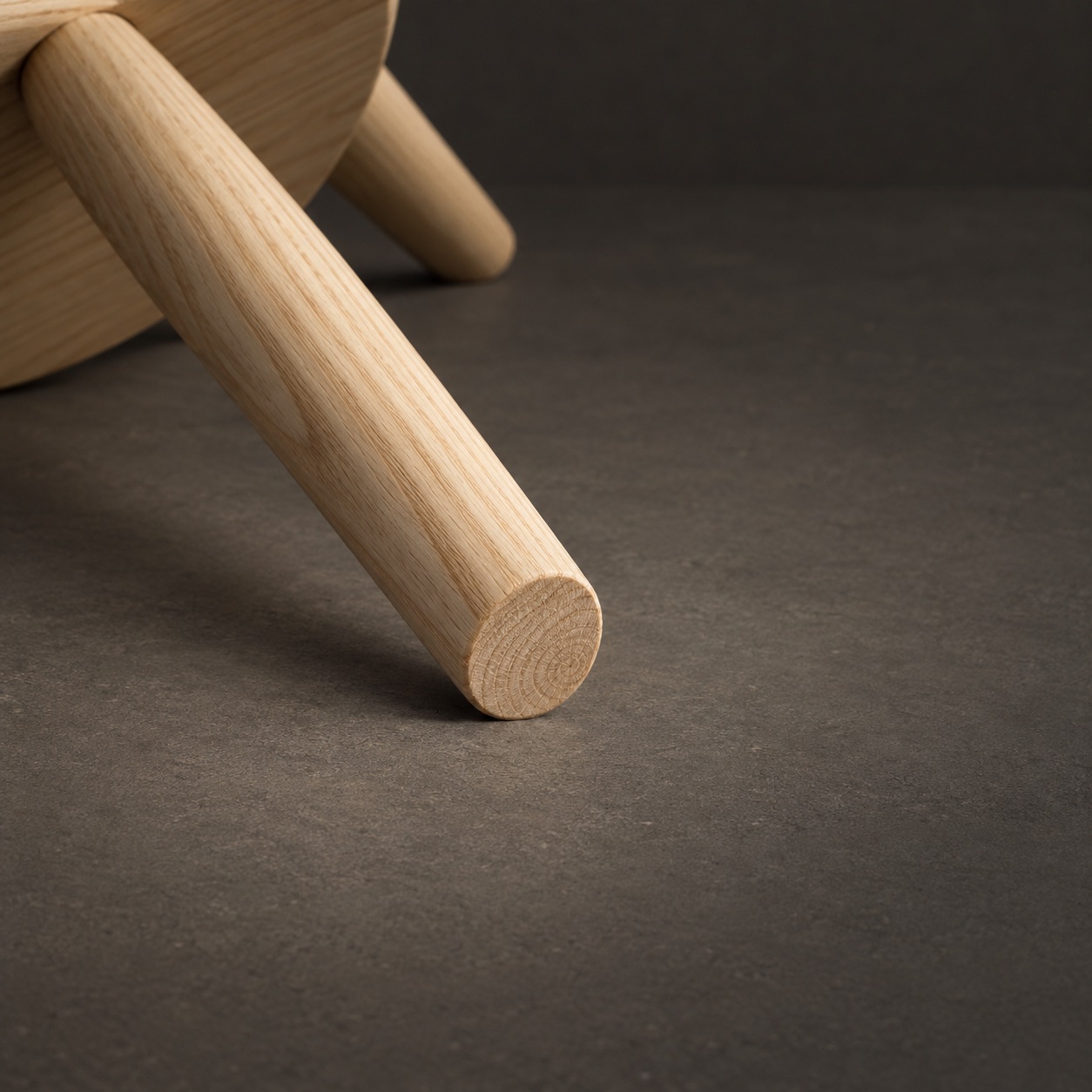 The stool laid on its side, showing the flat oiled end grain of one foot resting on the graphite floor.