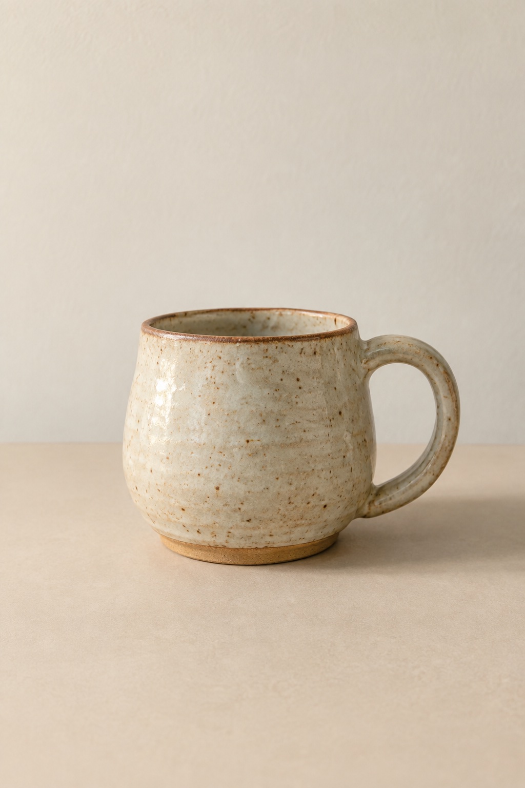 A mug in a speckled oatmeal glaze with a thin rust rim, alone on a warm matte surface in soft morning light.