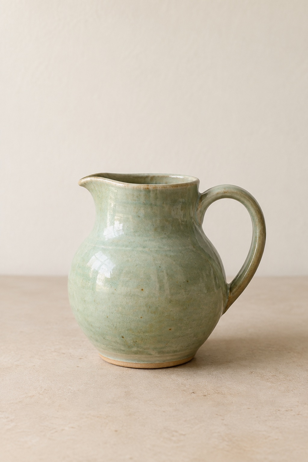 A small jug in a pale green celadon glaze, alone on a warm matte surface in soft morning light.