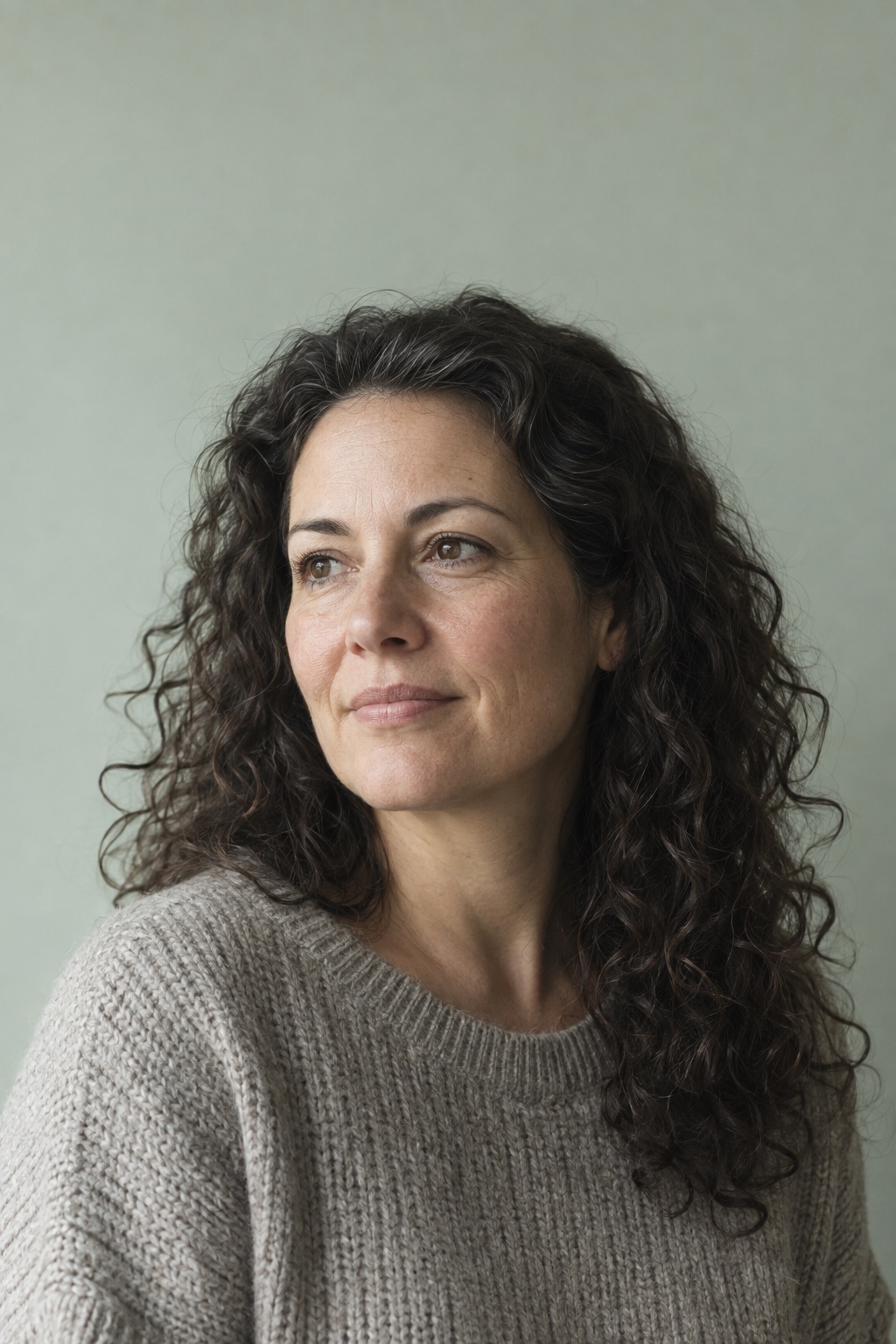 Portrait of a woman with long dark curly hair in a grey knitted jumper, turned towards a window and looking just past the camera.