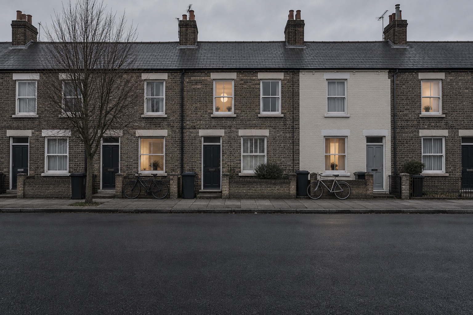 A terraced street at dusk seen square-on from across the road: brick and painted house fronts, dark bins, two bicycles and a few lit windows above an empty road.