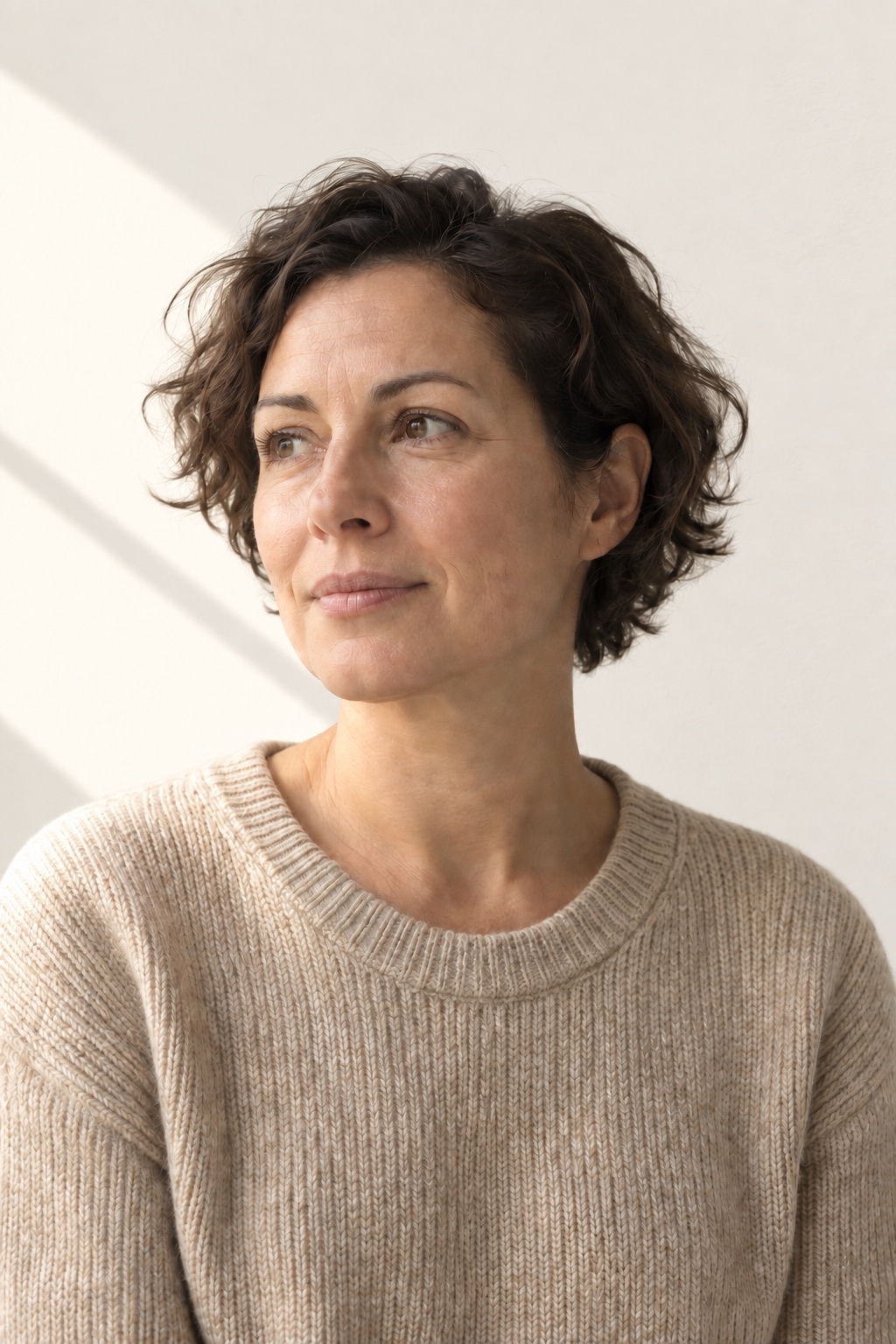 A research editor with short dark curly hair in an oatmeal sweater, late window light on the wall behind her.